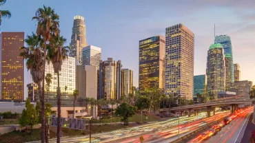 Los Angeles skyline at dusk with illuminated buildings and palm trees.