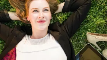 Woman lying on grass, smiling, with shoes and tablet nearby.