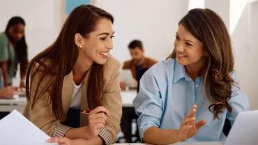 Two women smiling and talking while working on a laptop in an office setting.