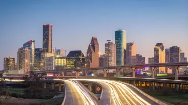 City skyline at dusk with illuminated skyscrapers and light trails on the highway.