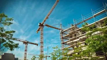 Three cranes towering over a building under construction with scaffolding against a blue sky.